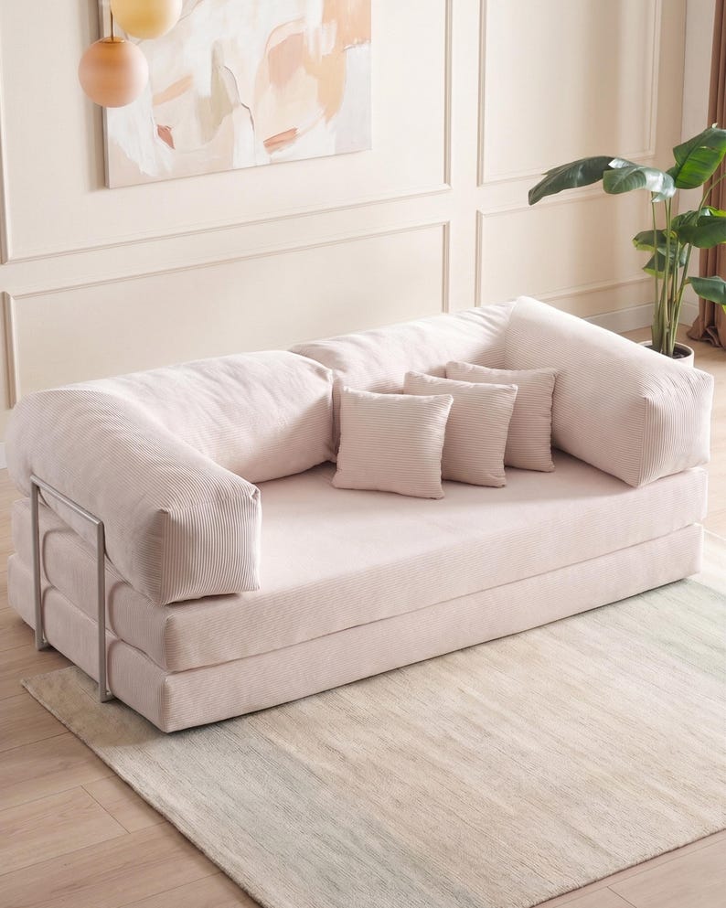 May include: A light pink, corduroy-textured convertible sofa with three matching throw pillows. The sofa has a metal frame and is set on a light-colored rug. The room has a neutral color palette with a painting and a potted plant.
