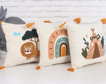 May include: Three off-white decorative throw pillows with embroidered designs. One features a lion, tree, and cloud. Another has a rainbow, and the third displays a teepee. Each pillow has orange tassels at the corners.