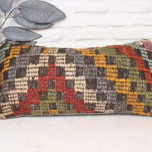 Puede incluir: Cojín decorativo rectangular con un patrón geométrico tejido en tonos marrón, rojo, amarillo, verde y beige. El cojín está colocado sobre una superficie blanca, con una planta en una maceta decorativa en el fondo.