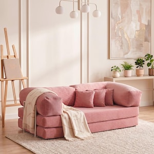May include: A pink corduroy sofa with two matching throw pillows and a cream-colored blanket with a heart design. The sofa sits on a beige rug in a room with a painting and plants.