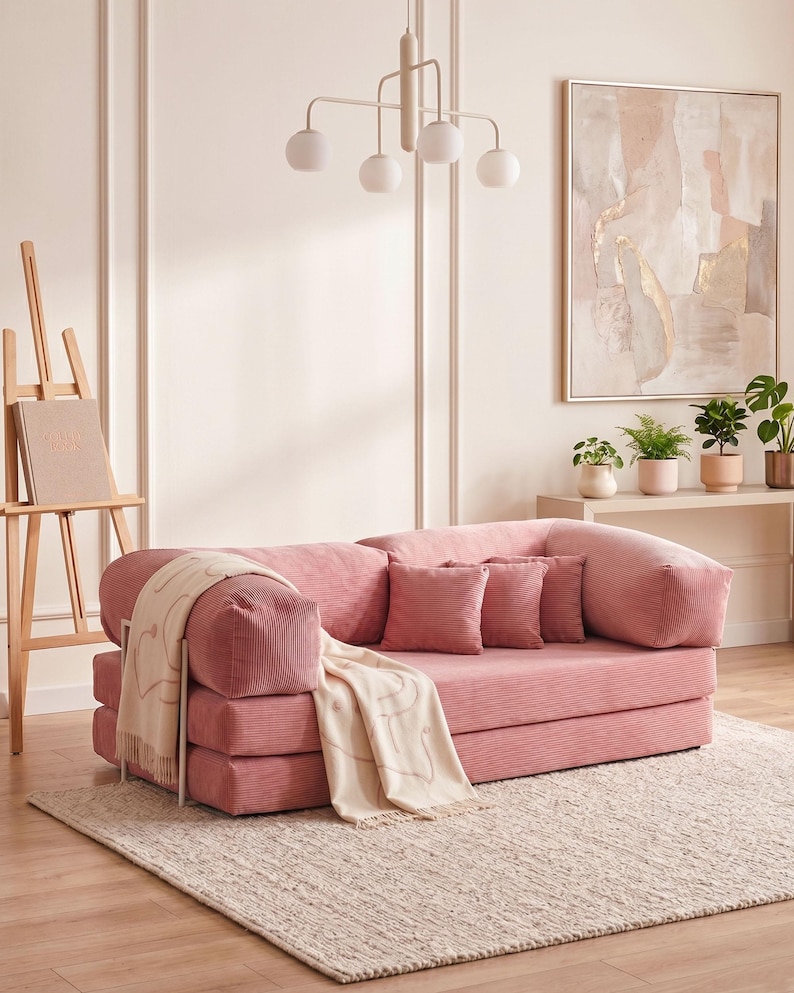May include: A pink corduroy sofa with two matching throw pillows and a cream-colored blanket with a heart design. The sofa sits on a beige rug in a room with a painting and plants.