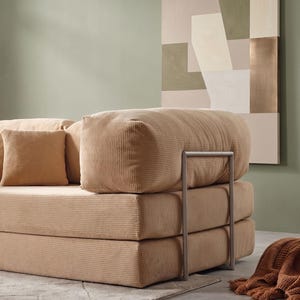 May include: A beige corduroy sofa with a stacked design, featuring a square armrest and matching pillows. The sofa has a metal support frame and sits on a patterned rug. A painting with geometric shapes hangs on the wall.