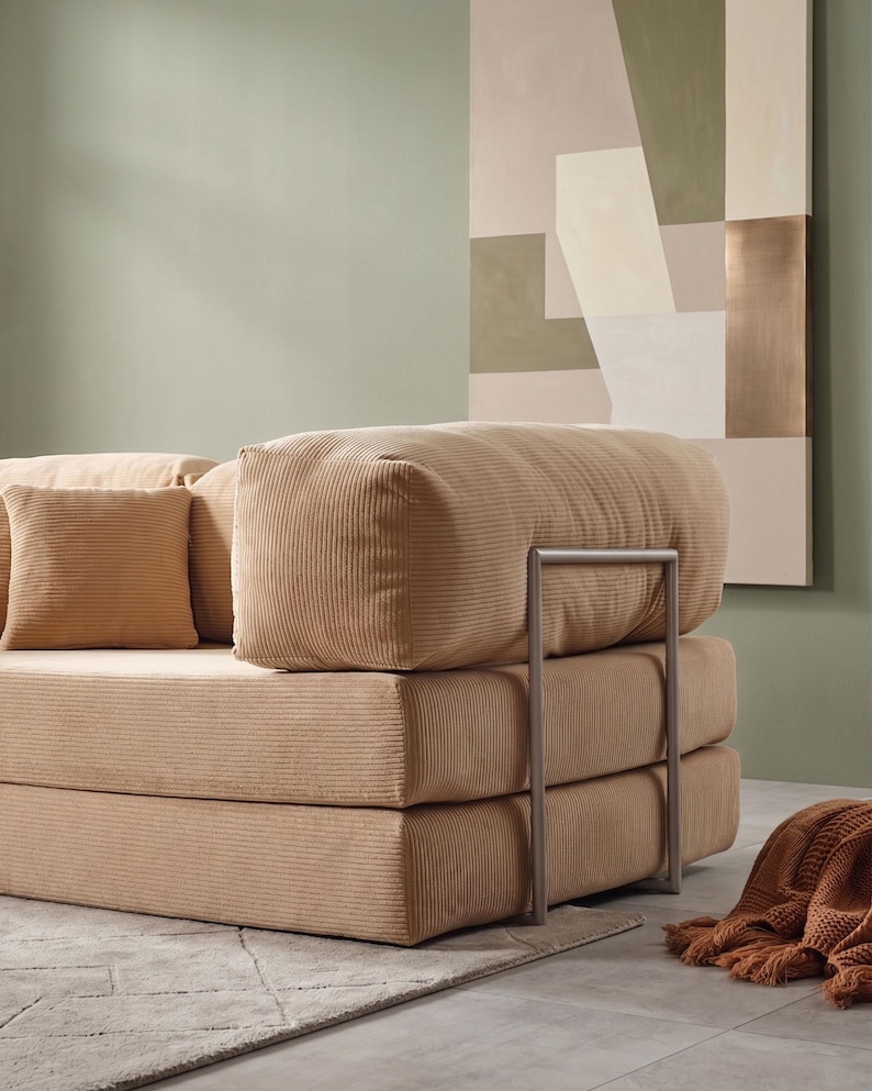 May include: A beige corduroy sofa with a stacked design, featuring a square armrest and matching pillows. The sofa has a metal support frame and sits on a patterned rug. A painting with geometric shapes hangs on the wall.