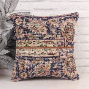 May include: Square decorative pillow with a vintage rug design. The pillow features a navy blue background with floral patterns in shades of beige, pink, and green. A brown leather strip accents the center. The pillow is placed against a white brick wall.