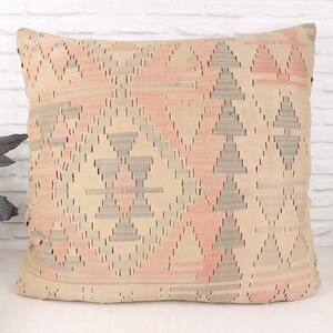 May include: A square decorative pillow with a geometric pattern in beige, pink, and gray. The pillow features a repeating design of triangles and diamond shapes, creating a textured look. The pillow is placed against a white brick wall.