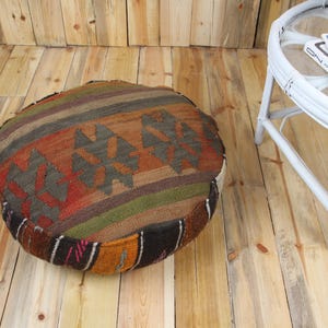 Pode incluir: Almofada de chão redonda com um padrão geométrico colorido. A almofada apresenta uma mistura de listras e formas marrons, laranja, verdes e pretas. A almofada está colocada em um chão de madeira.