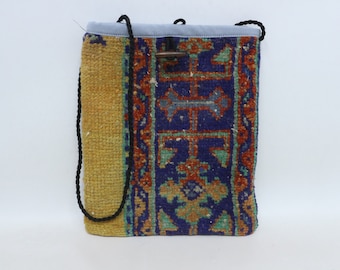 May include: A rectangular shoulder bag with a woven rug design. The bag features a blue top, a wooden toggle closure, and a black cord strap. The rug pattern includes shades of blue, orange, and green on a dark background, with a yellow side panel.