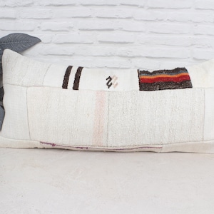 Pode incluir: Almofada retangular branca com design de patchwork. Apresenta detalhes em castanho, vermelho e laranja. A almofada é feita de um tecido texturizado e tem uma paleta de cores neutras, adequada para vários estilos de decoração.