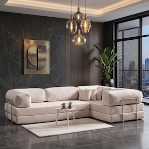 May include: A modern living room featuring a large, light pink sectional sofa with a matching rug. Above, a cluster of glass pendant lights hangs from the ceiling. A small side table holds a cup and plant. The room has dark gray flooring and a large window.