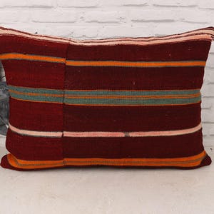 May include: A rectangular cushion with a burgundy base and horizontal stripes in orange, teal, and pink. The cushion has a textured appearance and is likely made of woven fabric. The cushion is against a white brick wall.