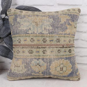 Può includere: Cuscino quadrato decorativo dall'aspetto vintage e usurato. Il cuscino presenta un design tessuto nei toni del beige, blu e marrone. Il motivo include elementi floreali e geometrici, creando un aspetto strutturato. Il cuscino è posto davanti a un muro di mattoni bianchi.