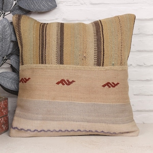 May include: Square decorative pillow with a striped top section in shades of tan and brown. The lower section is tan with three red decorative elements. The bottom edge has a purple border. The pillow is on a white brick background.