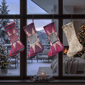 Peut inclure: Quatre chaussettes de Noël suspendues à une fenêtre. Trois chaussettes présentent un motif d'étoile géométrique magenta et rose avec des accents sombres. La quatrième est de couleur crème avec un imprimé végétal brun répétitif.
