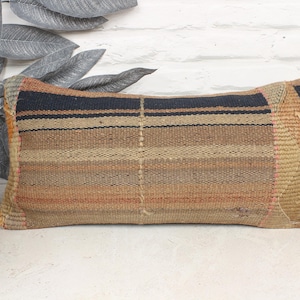 May include: Rectangular woven cushion with a striped pattern in shades of brown, tan, and black. The cushion has a textured appearance and is placed against a white brick wall. The cushion is approximately 50 cm long.