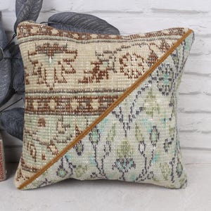 May include: Square decorative pillow with a patchwork design. The pillow features a beige and brown patterned top half and a light green and blue patterned bottom half, divided by a diagonal brown seam. The pillow is placed in front of a white brick wall.