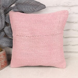 May include: A square, light pink woven pillow with a decorative stitched detail. The pillow is placed in front of a white brick wall and next to a decorative plant in a square pot.