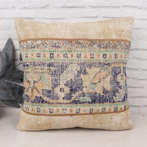 May include: Square beige pillow with a central patterned design in navy blue, teal, and tan. The pillow has a textured appearance, suggesting a woven or rug-like material, and is likely used for home decor.