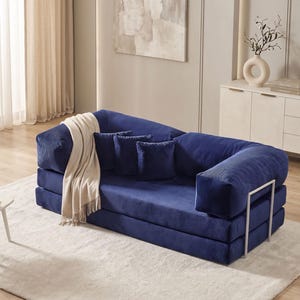 May include: A modern, modular blue corduroy sofa with three matching throw pillows and a cream-colored blanket. The sofa has a unique design with a metal frame and is placed on a light-colored rug. The room has neutral-toned walls and decor.