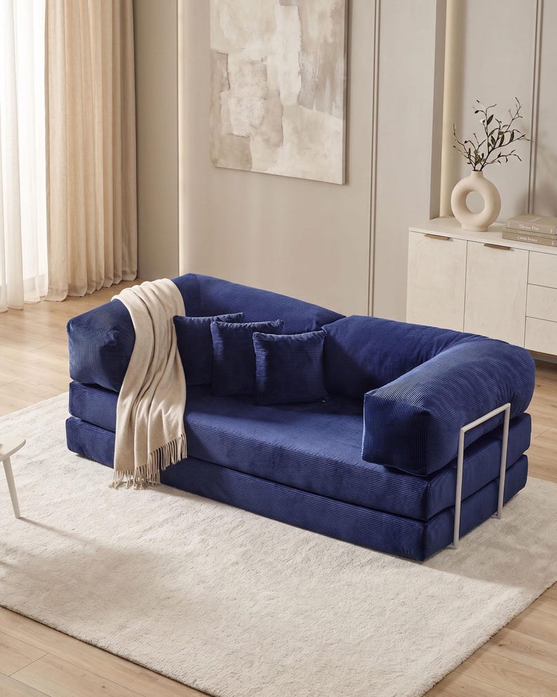 May include: A modern, modular blue corduroy sofa with three matching throw pillows and a cream-colored blanket. The sofa has a unique design with a metal frame and is placed on a light-colored rug. The room has neutral-toned walls and decor.