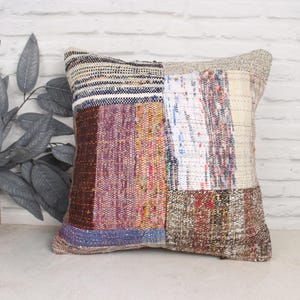 Può includere: Una federa patchwork con una varietà di colori e trame, tra cui marrone, bianco, blu e rosso. La federa è realizzata in tessuto tessuto e ha una forma quadrata.