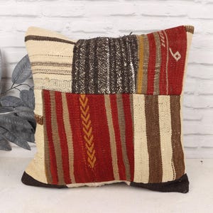 May include: A square decorative pillow with a patchwork design. The pillow features a mix of colors including cream, brown, red, and black. The pillow has a textured appearance and is likely made of woven fabric.