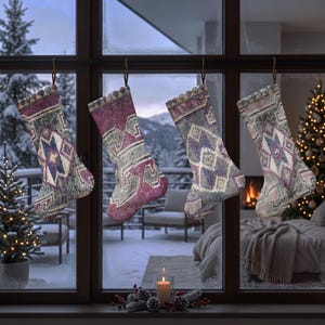 Puede incluir: Cuatro calcetines navideños colgados en fila frente a una ventana. Cada calcetín presenta un patrón geométrico tejido multicolor en tonos morado oscuro, verde apagado y crema, suspendidos por lazos marrones de una barra horizontal.