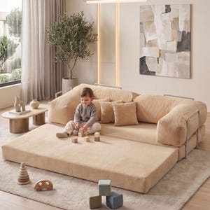 May include: A beige modular sofa with a corduroy texture, featuring a low profile and rounded edges. The sofa is accompanied by matching throw pillows. A child is playing with wooden blocks on the sofa. A wooden toy car and a stacking toy are on the floor.