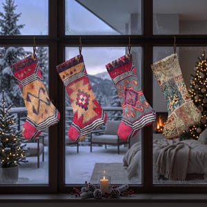 Puede incluir: Cuatro calcetines navideños colgados en fila frente a una ventana. Tres calcetines presentan patrones geométricos en rojo, amarillo y azul, mientras que el cuarto tiene un diseño floral y de cachemira multicolor. Una vela encendida está en el alféizar.