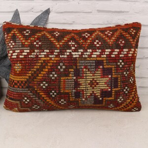 May include: Rectangular pillow with a woven, geometric pattern in shades of red, brown, orange, and white. The pillow has a textured appearance and is placed on a white surface.