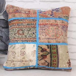 May include: Square patchwork pillow with a mix of patterned fabrics in beige, red, and blue. The pillow is divided into four sections with blue stitching. The pillow is placed in front of a white brick wall.