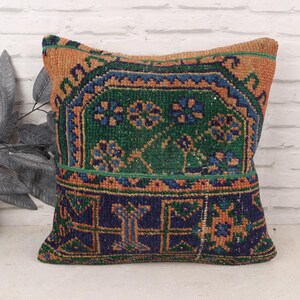 May include: Square decorative pillow with a traditional rug design. The pillow features a green, blue, and orange pattern. The pillow is placed in front of a white brick wall.