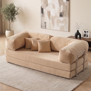 Puede incluir: Un sofá de pana beige con tres cojines a juego. El sofá tiene reposabrazos redondeados y un perfil bajo. La habitación presenta una paleta de colores neutros, una gran pintura abstracta y un olivo decorativo en una maceta blanca.