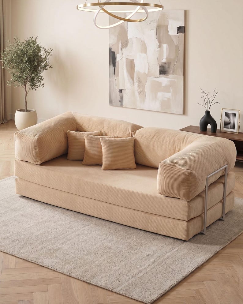 May include: A beige corduroy sofa with three matching throw pillows. The sofa has rounded armrests and a low profile. The room features a neutral color palette, a large abstract painting, and a decorative olive tree in a white pot.