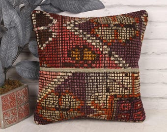 Puede incluir: Cojín decorativo cuadrado con un patrón geométrico tejido en tonos de rojo, marrón, naranja y morado. El cojín tiene un aspecto texturizado y una banda de color neutro en el medio. El cojín está colocado cerca de una planta decorativa.