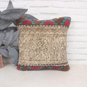 Op de afbeelding: Een decoratief kussen met een beige achtergrond en een kleurrijk geometrisch patroon rond de randen. Het patroon heeft tinten rood, blauw en bruin.