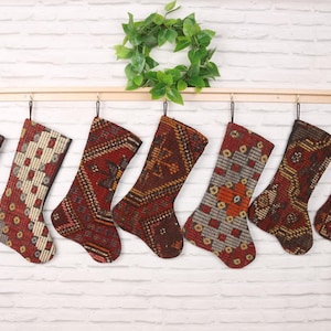 May include: Seven Christmas stockings with intricate patterns in shades of red, brown, and cream hang from a wooden rod. A green wreath sits above the stockings against a white brick wall. Each stocking has a unique design.