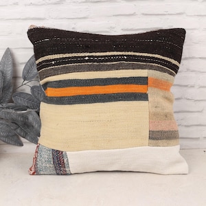 Puede incluir: Un cojín decorativo cuadrado con un diseño de patchwork. La parte superior es de color marrón oscuro, con rayas horizontales grises, naranjas y beiges. La parte inferior es una mezcla de beige y blanco, con una sección estampada en la base. El cojín está sobre una superficie blanca.