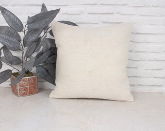 May include: A plain white pillow with a linen texture, sitting on a white brick wall background. A potted plant with gray leaves is in the foreground.