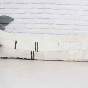 Puede incluir: Un cojín rectangular blanco roto con un diseño de patchwork. El cojín presenta rayas verticales negras y una superficie texturizada. Se coloca sobre una superficie blanca, con una planta decorativa en el fondo.