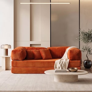 May include: An orange corduroy sofa with matching throw pillows and a cream-colored blanket. A round, light-colored coffee table holds books and a gold bowl. A small side table with a lamp and a potted plant complete the living room scene.