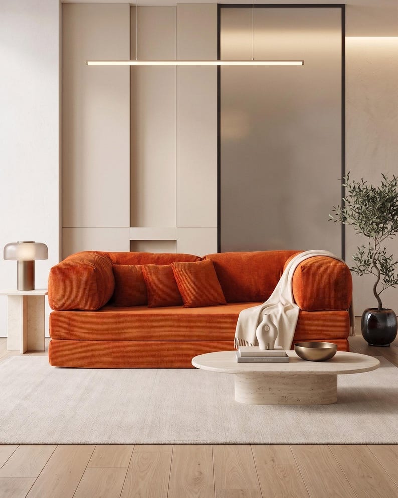 May include: An orange corduroy sofa with matching throw pillows and a cream-colored blanket. A round, light-colored coffee table holds books and a gold bowl. A small side table with a lamp and a potted plant complete the living room scene.