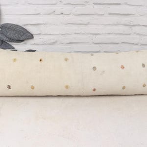 Peut inclure: Un long coussin rectangulaire crème avec des motifs floraux brodés de différentes couleurs. Le coussin est posé sur une surface blanche, avec un mur de briques en arrière-plan. Des feuilles décoratives et un petit pot sont visibles à gauche.