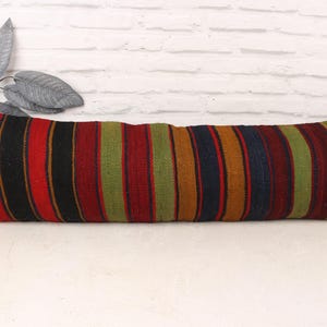 May include: A long, rectangular pillow with a striped pattern in shades of red, black, green, and brown. The pillow is placed on a white surface against a white brick wall. The pillow's dimensions are not visible.