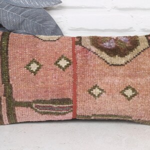 May include: Rectangular decorative pillow with a vintage rug design. The pillow features a pink and brown color scheme with geometric patterns and a central vertical red stripe. The edges are trimmed in a darker brown.