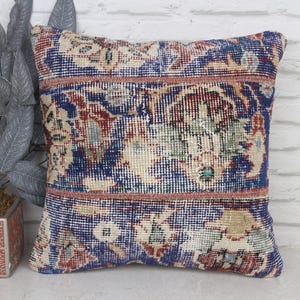 May include: Square decorative pillow with a vintage rug design. The pillow features a blue, red, and beige floral pattern with a distressed look. The pillow is placed in front of a white brick wall.