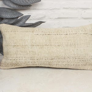 May include: A rectangular beige pillow with a woven texture. The pillow has a neutral color palette and is placed against a white brick wall. The pillow is approximately 12 inches wide and 20 inches long.