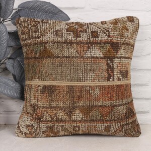 May include: Square decorative pillow with a woven, textured design in shades of brown, beige, and orange. The pillow features a geometric pattern and a neutral-colored band across the middle. The pillow is placed in front of a white brick wall.