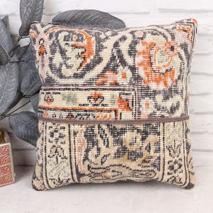May include: Square decorative pillow with a patchwork design. The pillow features a mix of colors, including orange, black, and beige, with a vintage-inspired pattern. The pillow is placed in front of a white brick wall.
