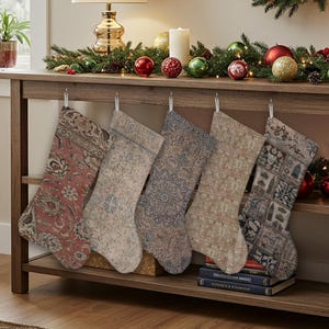 May include: Five Christmas stockings with vintage rug patterns hang from a wooden shelf. The stockings feature various colors, including red, blue, and beige, and are decorated with traditional floral and geometric designs. A festive garland with ornaments and lights adorns the shelf.