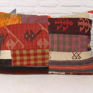 May include: Rectangular patchwork pillow with a variety of patterns and colors. The pillow features red, orange, burgundy, and beige patches with geometric and plaid designs. The pillow is placed on a white surface.